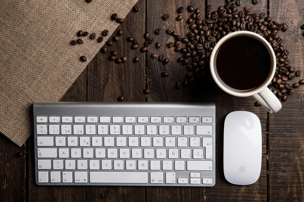 The image depicts a keyboard, mouse, and a cup of coffee on top of a dark wood desk. Coffee beans decorate the area around the cup of coffee.